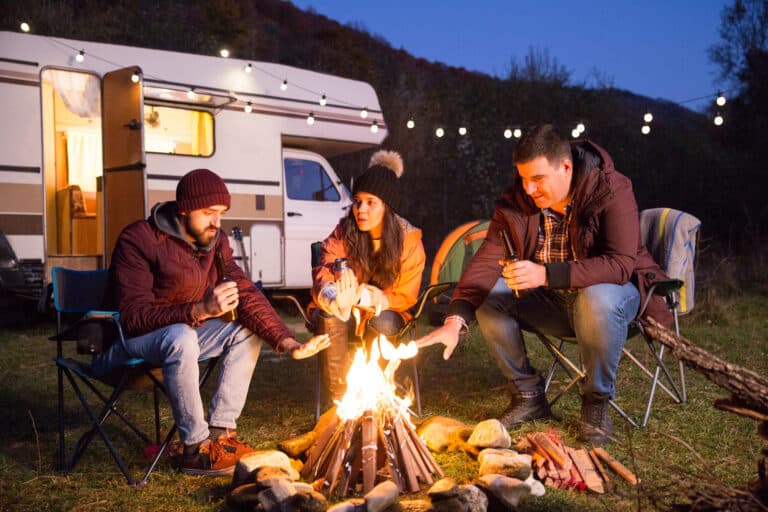 Three people sit in chairs around a campfire near a camper van, warming their hands. They are wearing warm clothing and appear to be in a wooded area during dusk.