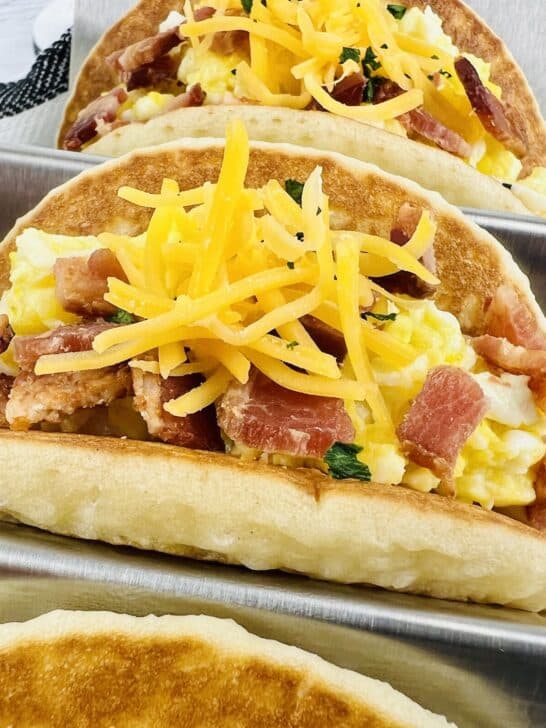 pancakes in stainless steel taco holders filled with scrambled eggs, shredded cheese and bacon with white and black striped towel in background