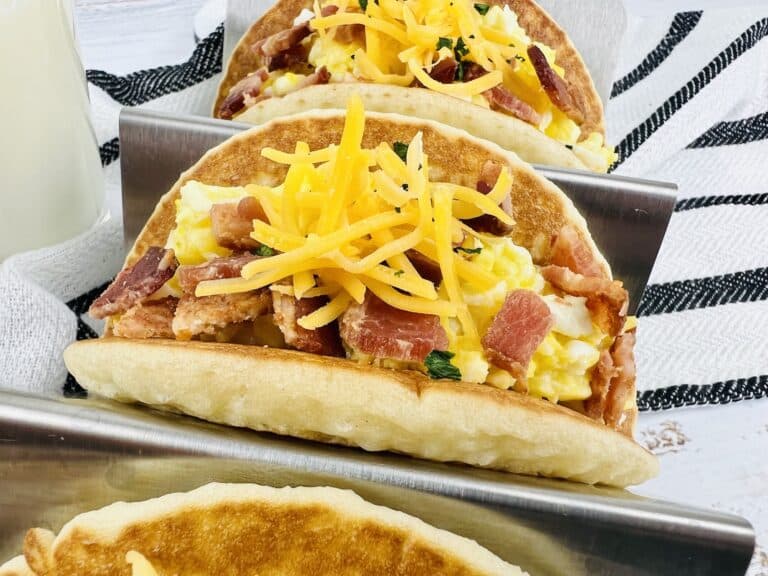 pancakes in stainless steel taco holders filled with scrambled eggs, shredded cheese and bacon with white and black striped towel in background