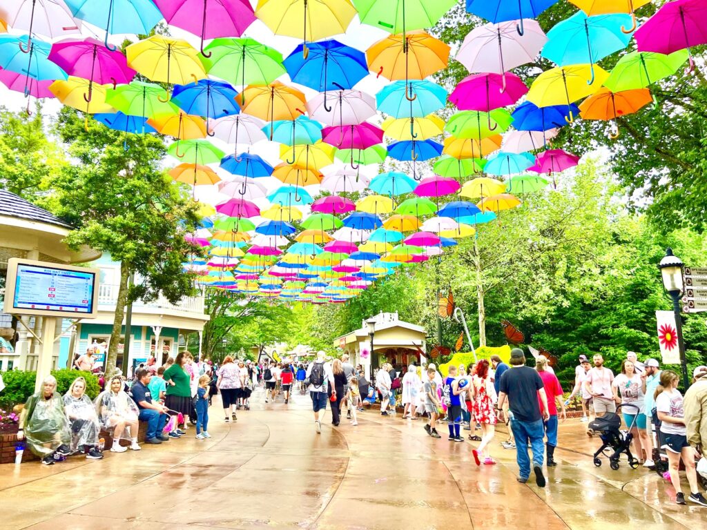 Visitors stroll through Dollywood's vibrant outdoor park, delighting in a colorful canopy of umbrellas that provides a whimsical touch amidst the surrounding trees and buildings.