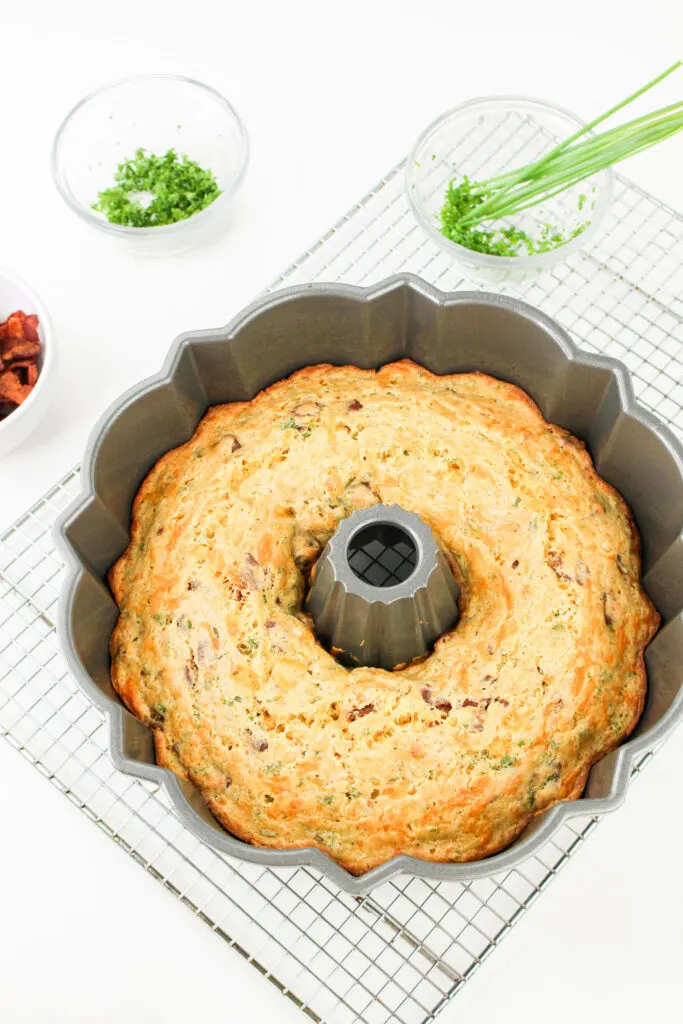 A baked savory bundt cake in a fluted pan on a cooling rack with dishes of chopped herbs and bacon bits nearby.