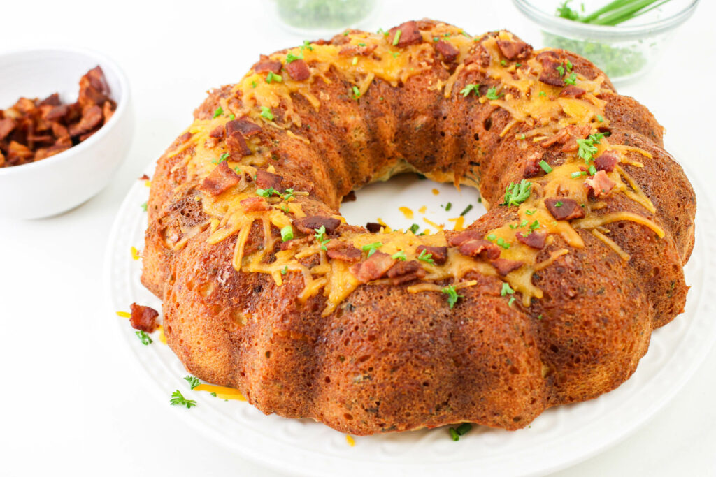 A savory bundt cake topped with bacon pieces and herbs on a white plate, with a bowl of bacon pieces and a glass bowl in the background.