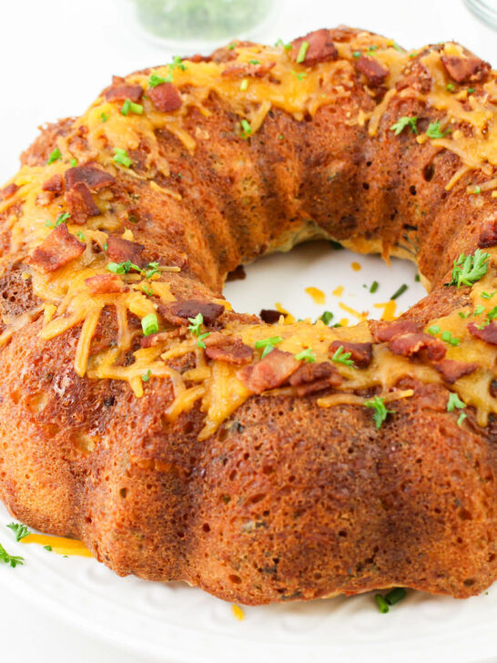 A savory bundt cake topped with bacon pieces and herbs on a white plate, with a bowl of bacon pieces and a glass bowl in the background.