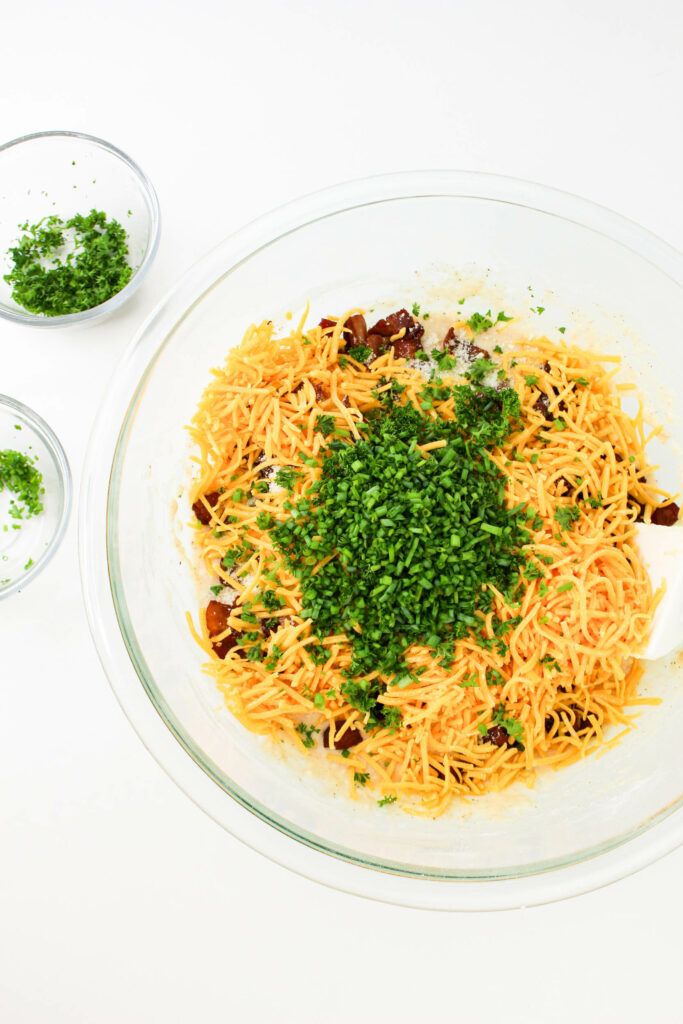 A clear glass bowl containing shredded cheese, chopped green herbs, and some other ingredients, with two smaller bowls of chopped herbs nearby on a white surface.