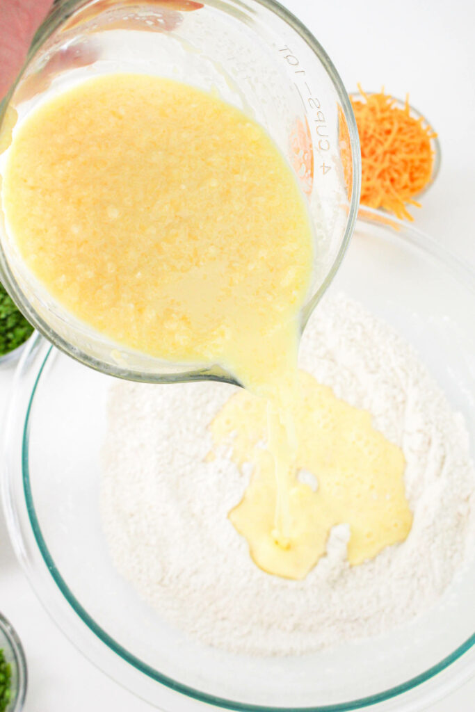 A hand pouring a yellow liquid mixture from a measuring cup into a bowl containing dry ingredients. Green onions and shredded cheese are present in the background.