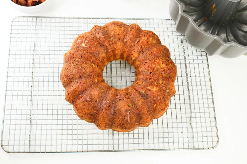 A baked Bundt cake sits on a cooling rack next to an empty Bundt pan on a white surface.