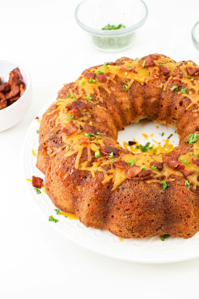 A Bundt-shaped bacon cheese bread garnished with herbs and placed on a white plate, surrounded by bowls of bacon pieces and herbs.