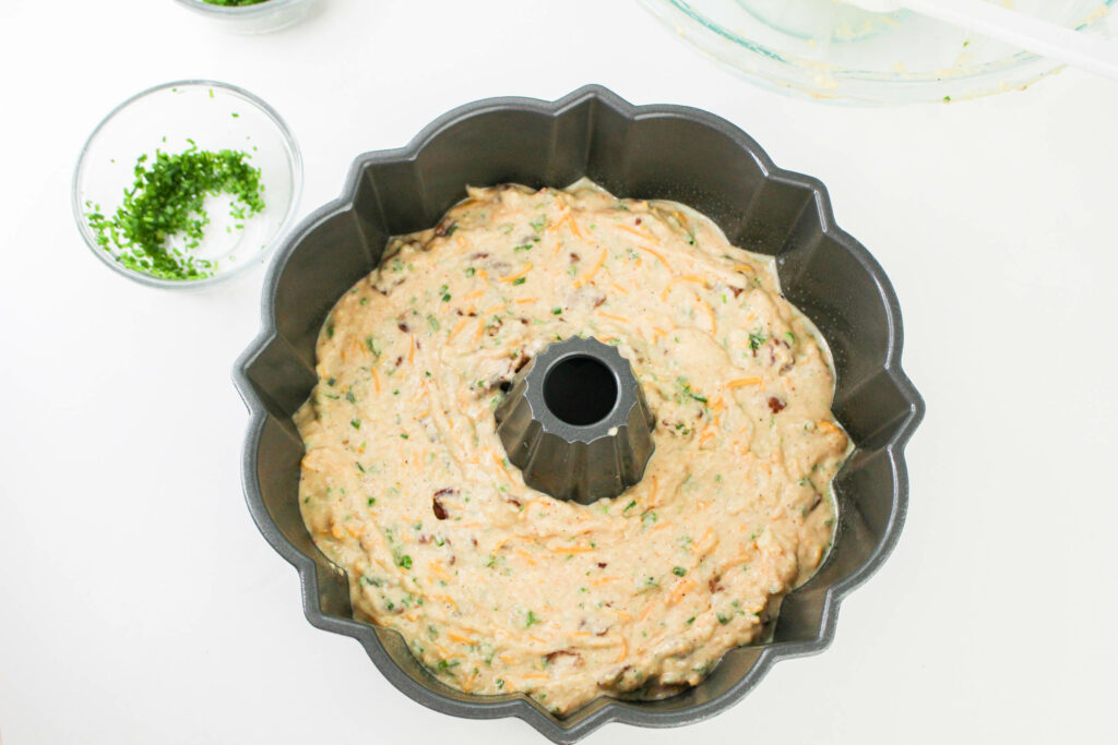 Bundt cake pan filled with raw batter containing visible herbs, placed on a white surface next to a small bowl of chopped herbs and a clear mixing bowl.