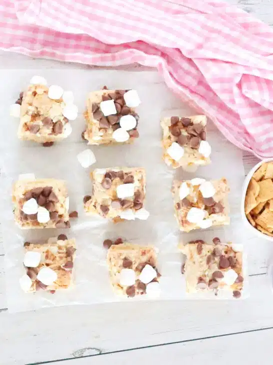 Overhead view of 12 square s'mores rice crispy treats with chocolate chips and marshmallows on parchment paper. Nearby are a pink cloth, a bowl of cereal, and a bowl of marshmallows on a wooden surface.