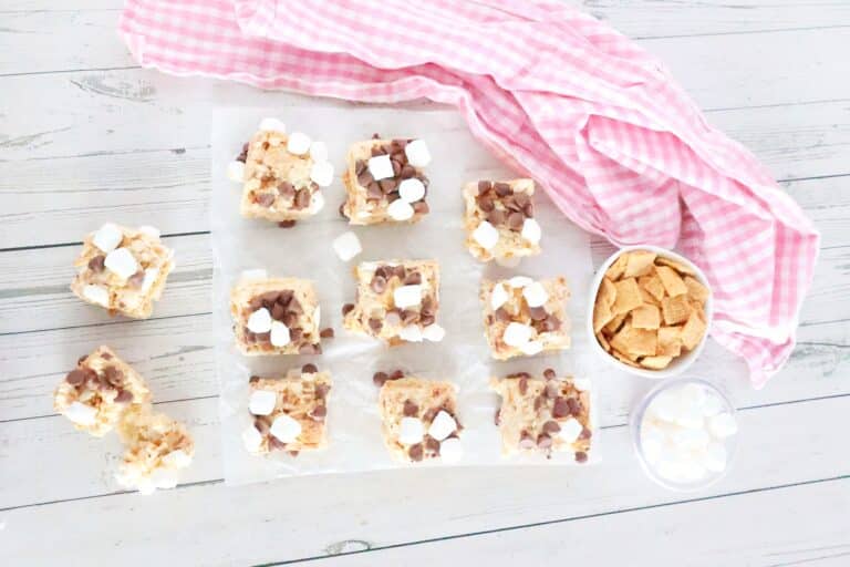Overhead view of 12 square s'mores rice crispy treats with chocolate chips and marshmallows on parchment paper. Nearby are a pink cloth, a bowl of cereal, and a bowl of marshmallows on a wooden surface.