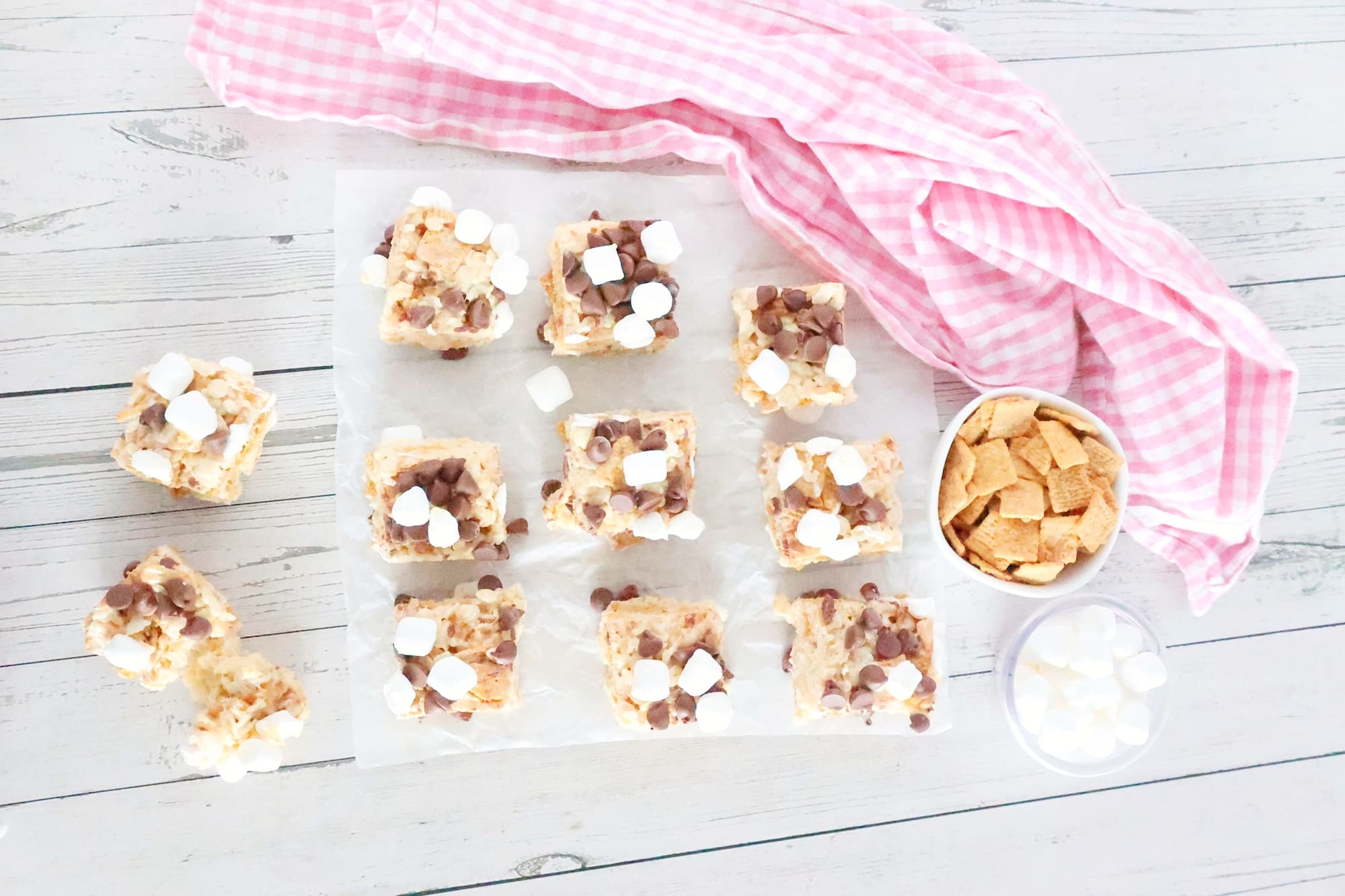 Overhead view of 12 square s'mores rice crispy treats with chocolate chips and marshmallows on parchment paper. Nearby are a pink cloth, a bowl of cereal, and a bowl of marshmallows on a wooden surface.