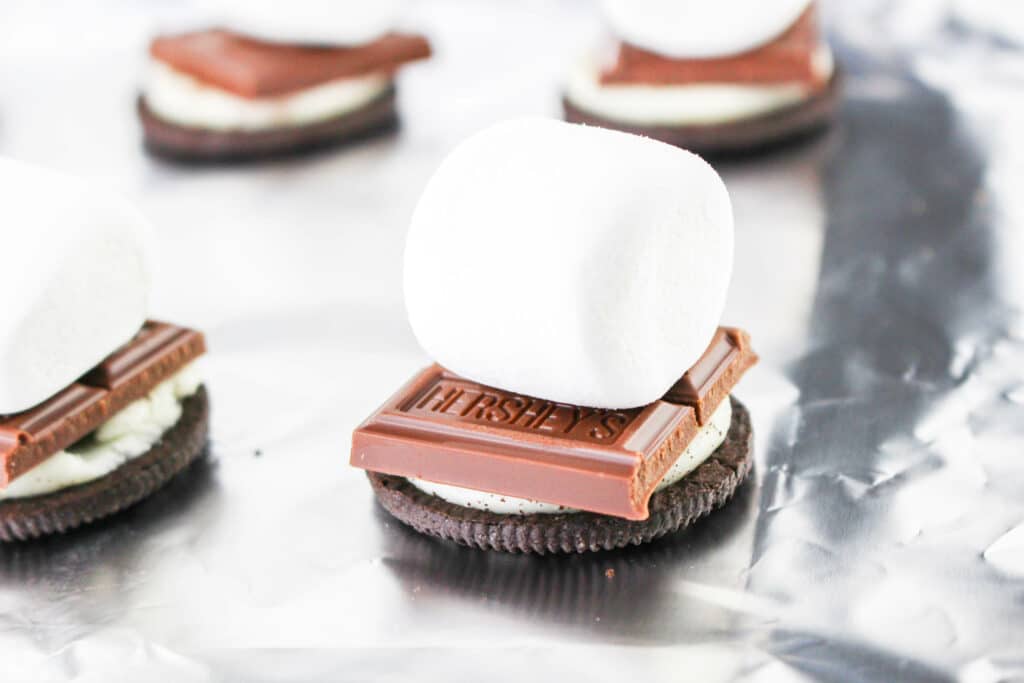 Close-up of an Oreo cookie topped with a square of chocolate and a marshmallow, resting on a foil surface.