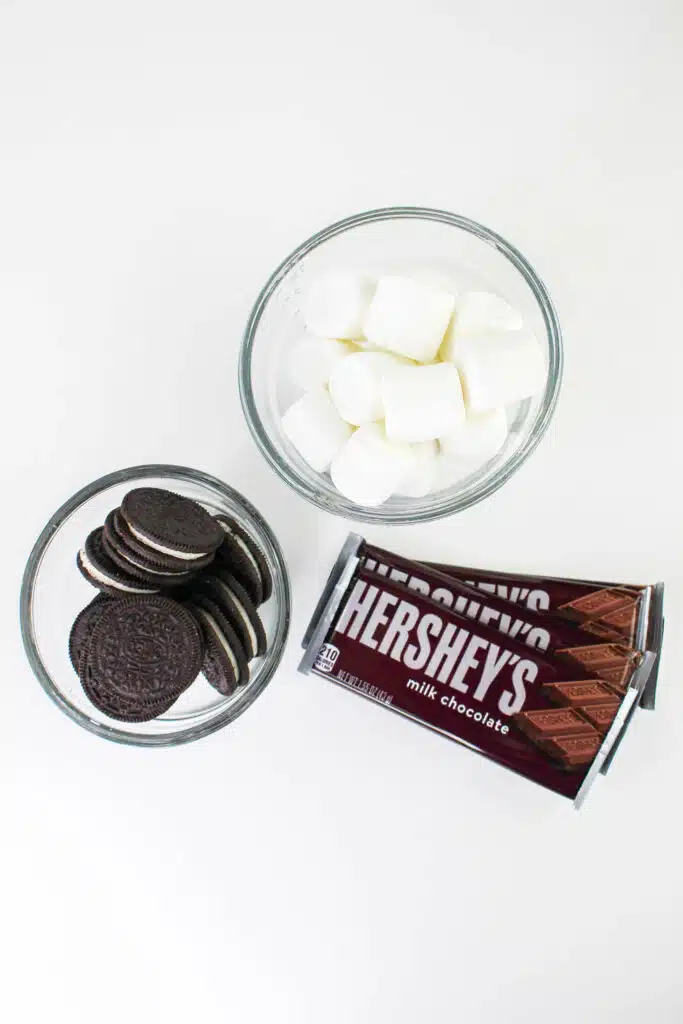 Two bowls filled with marshmallows and chocolate sandwich cookies next to Hershey's milk chocolate bars on a white surface.