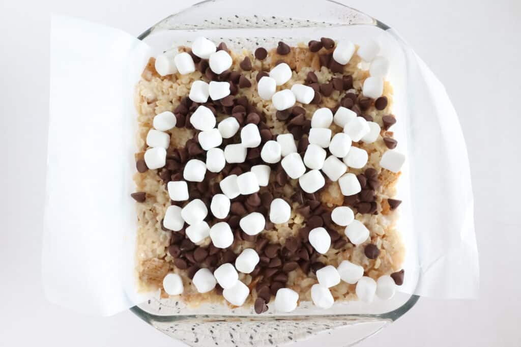 A glass dish with rice cereal treats topped with chocolate chips and mini marshmallows on parchment paper.