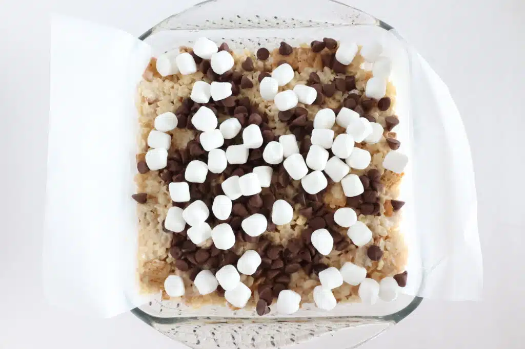 A glass dish with rice cereal treats topped with chocolate chips and mini marshmallows on parchment paper.