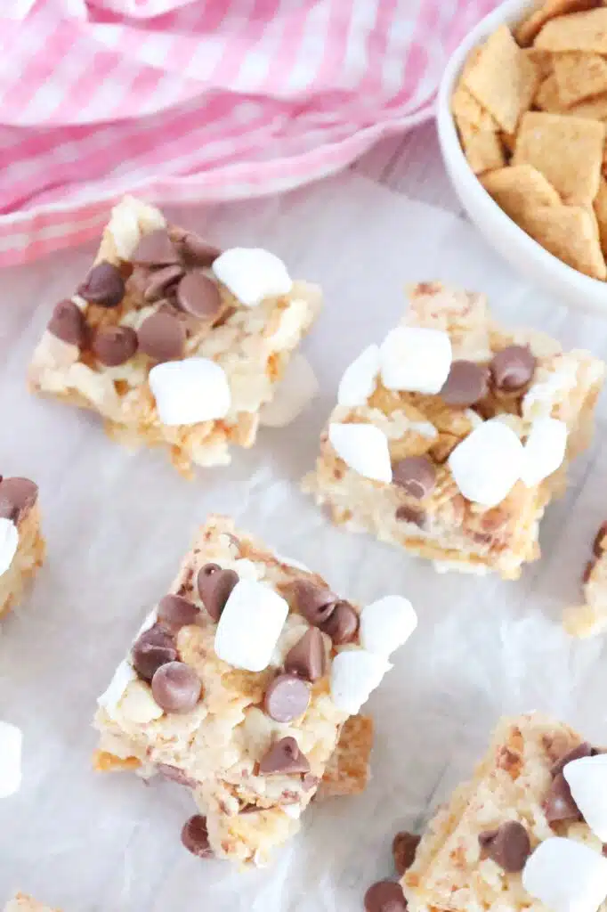 Squares of dessert bars topped with chocolate chips and mini marshmallows on parchment paper, with a bowl of cereal in the background.