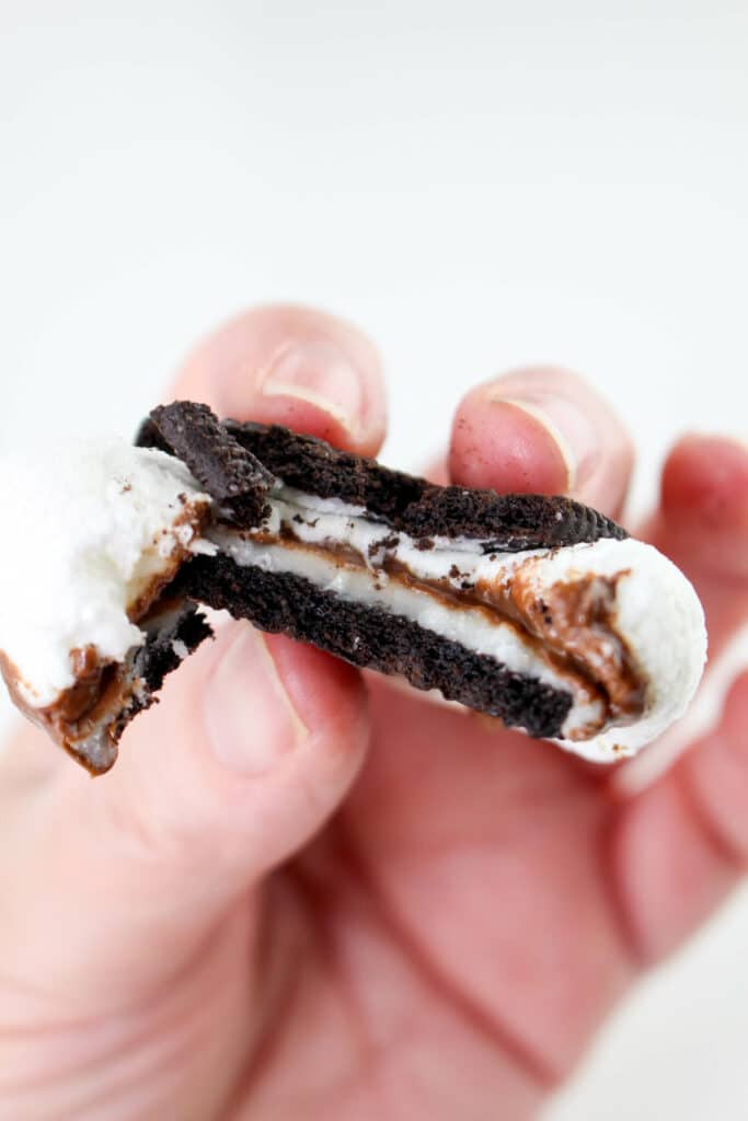 A hand holds a chocolate cookie with a marshmallow and chocolate filling, partially bitten.