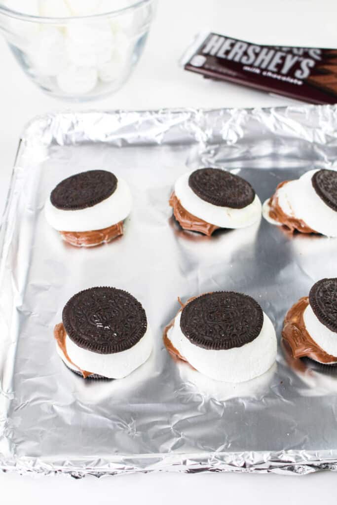 Oreo cookies with marshmallows and chocolate pieces, arranged on a foil-lined baking sheet.