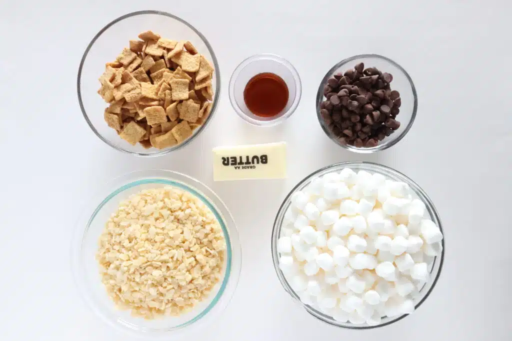 Ingredients for a sweet treat: cereal squares, vanilla, chocolate chips, butter, rice cereal, and mini marshmallows arranged in bowls on a white surface.