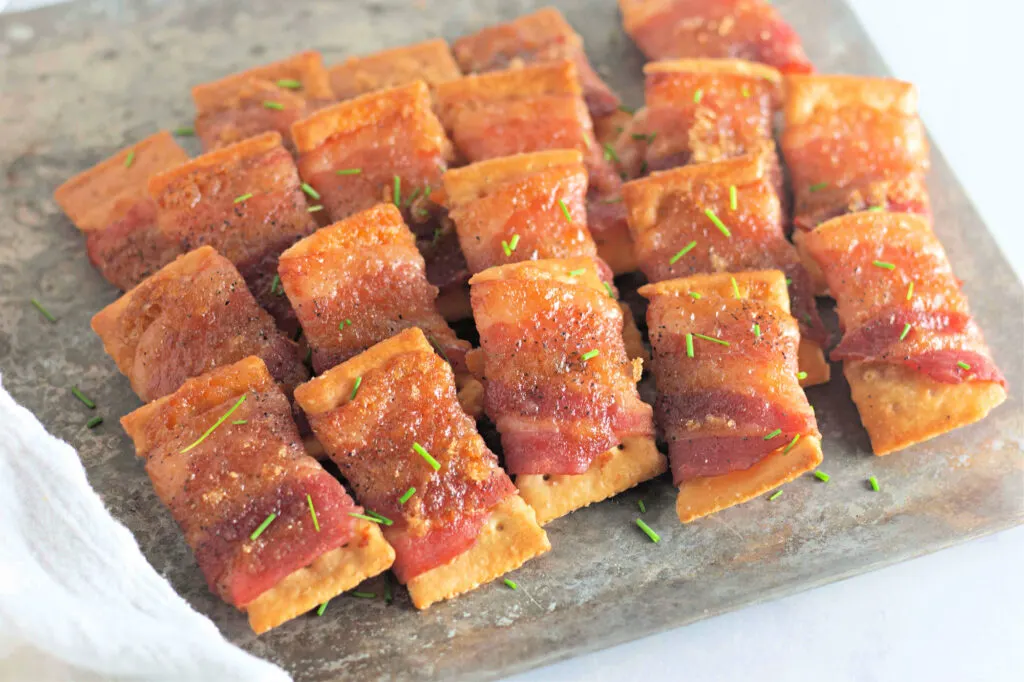 Bacon-wrapped crackers topped with herbs on a baking sheet.