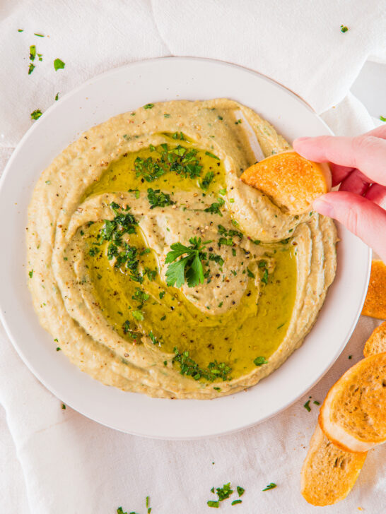 A bowl of hummus garnished with herbs and olive oil. A hand is dipping a slice of bread into the hummus.