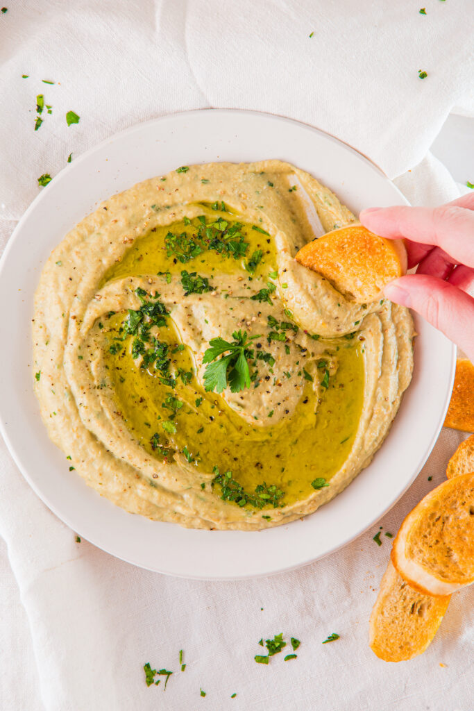 A bowl of hummus garnished with herbs and olive oil. A hand is dipping a slice of bread into the hummus.