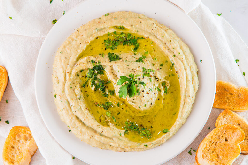 A plate of hummus garnished with olive oil and herbs, surrounded by slices of toasted bread on a white cloth.