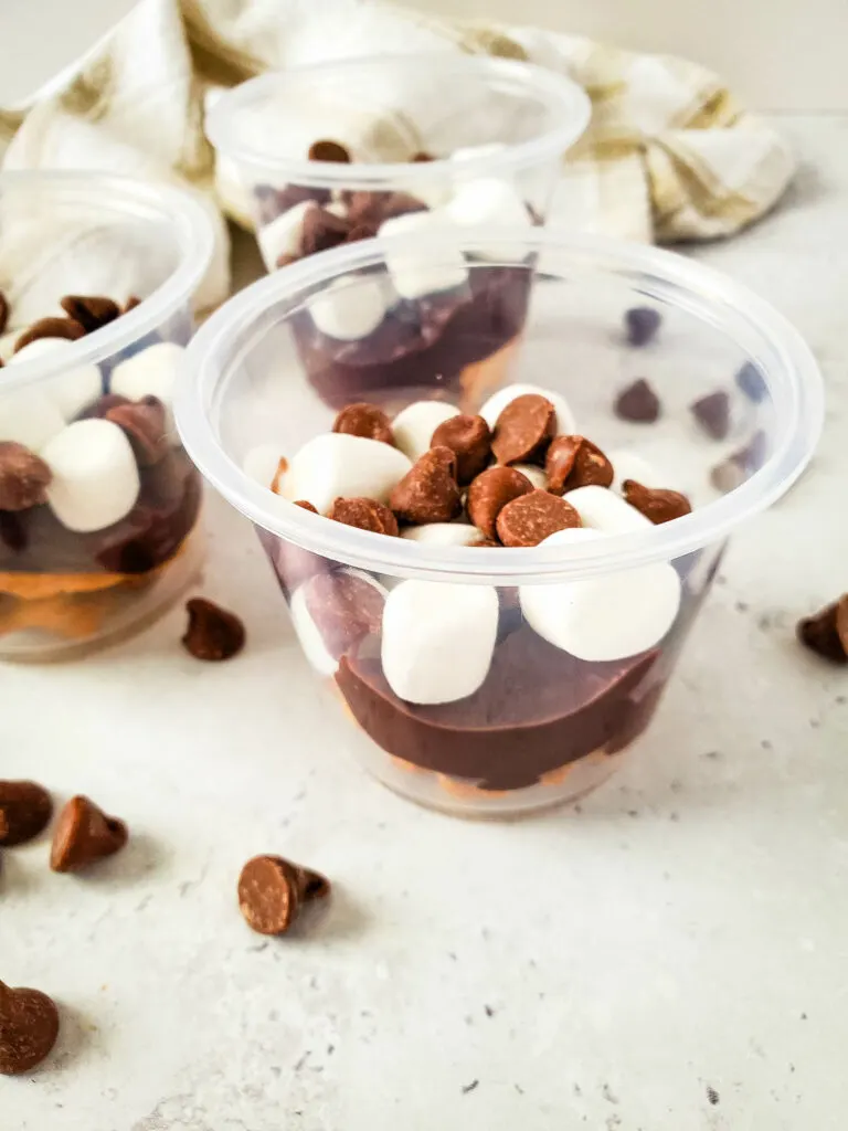 Three s'mores pudding cups filled with layers of chocolate chips and mini marshmallows rest on a light surface, with a checkered cloth in the background.