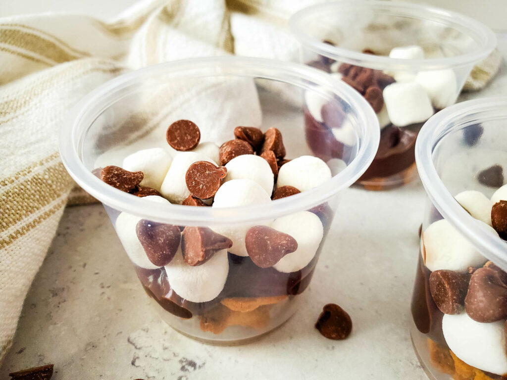 Clear plastic cups filled with layers of chocolate pudding, mini marshmallows, chocolate chips, and crushed graham crackers sit on the countertop. These delightful s'mores pudding cups capture all the campfire flavors without leaving your kitchen.