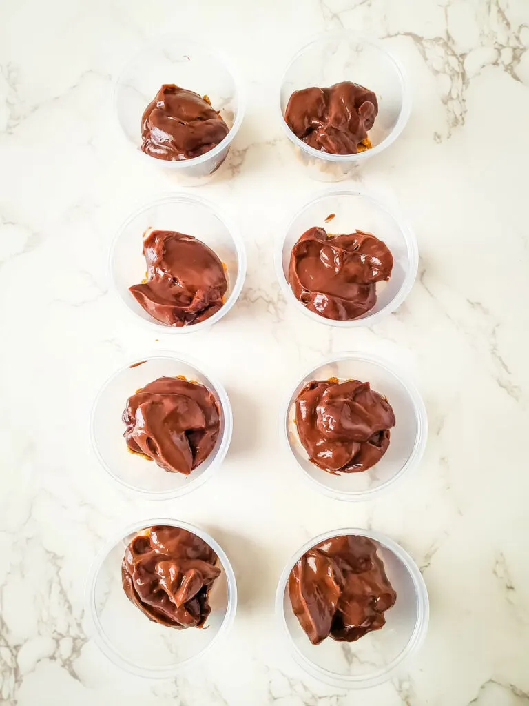 Eight plastic cups filled with chocolate-coated pastries, reminiscent of s'mores pudding cups, are arranged in two columns on a marble surface.