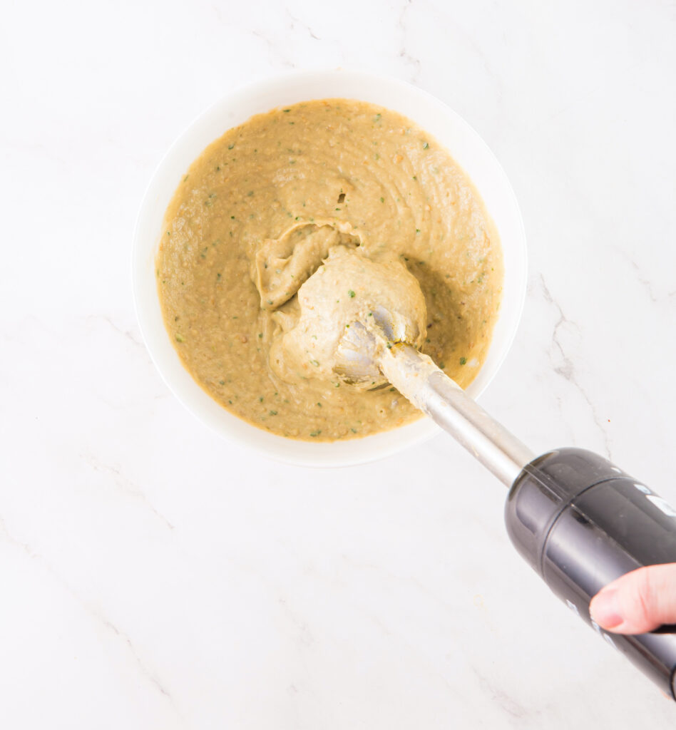 A hand uses an immersion blender to puree ingredients in a white bowl on a marble countertop.