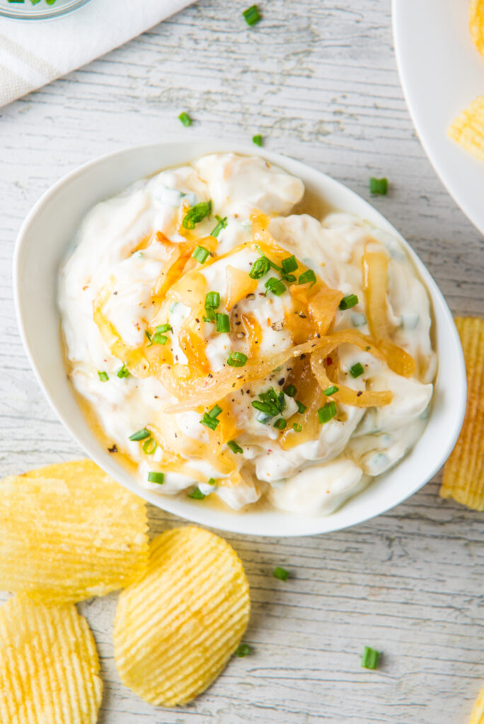 A bowl of French onion dip, creamy and topped with chopped chives and caramelized onions, sits invitingly on a wooden surface, surrounded by crispy potato chips.