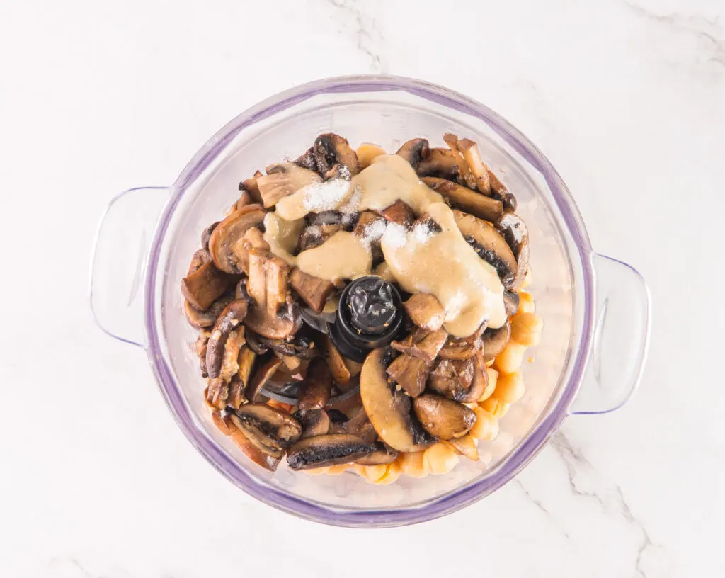 Overhead view of mushrooms, chickpeas, and sauce in a food processor on a marble surface, ready for blending into a delightful mushroom hummus.