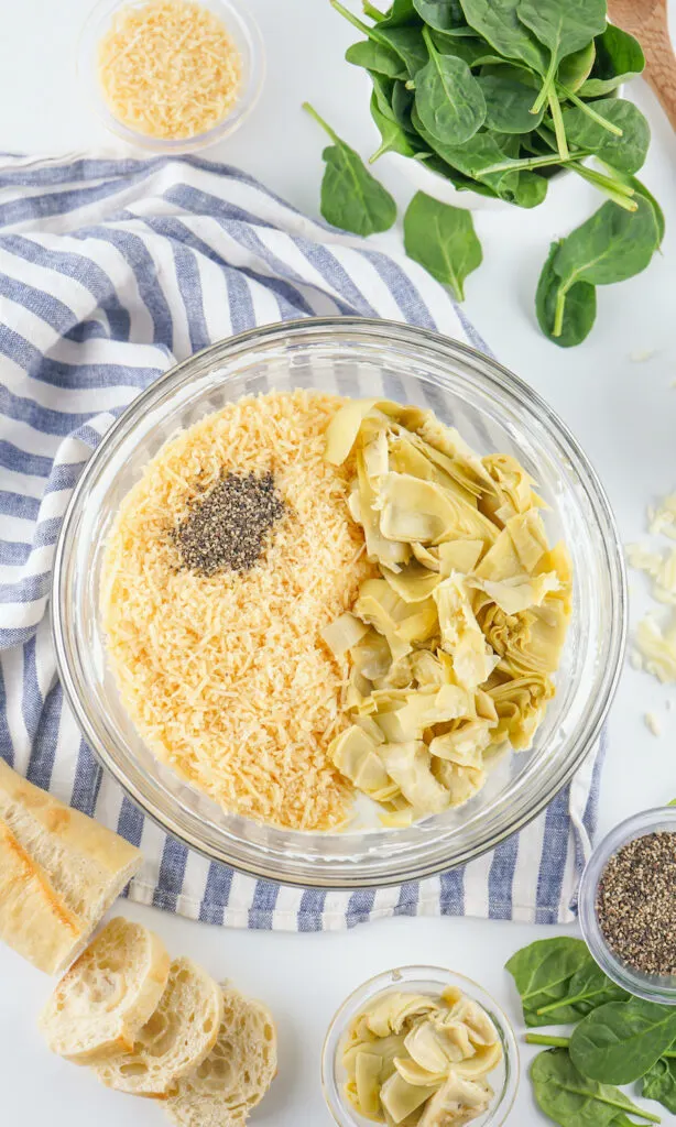 A glass bowl contains shredded cheese, chopped artichokes, and pepper. Surrounding it are spinach leaves, sliced bread, a striped cloth, and bowls of artichokes and pepper.