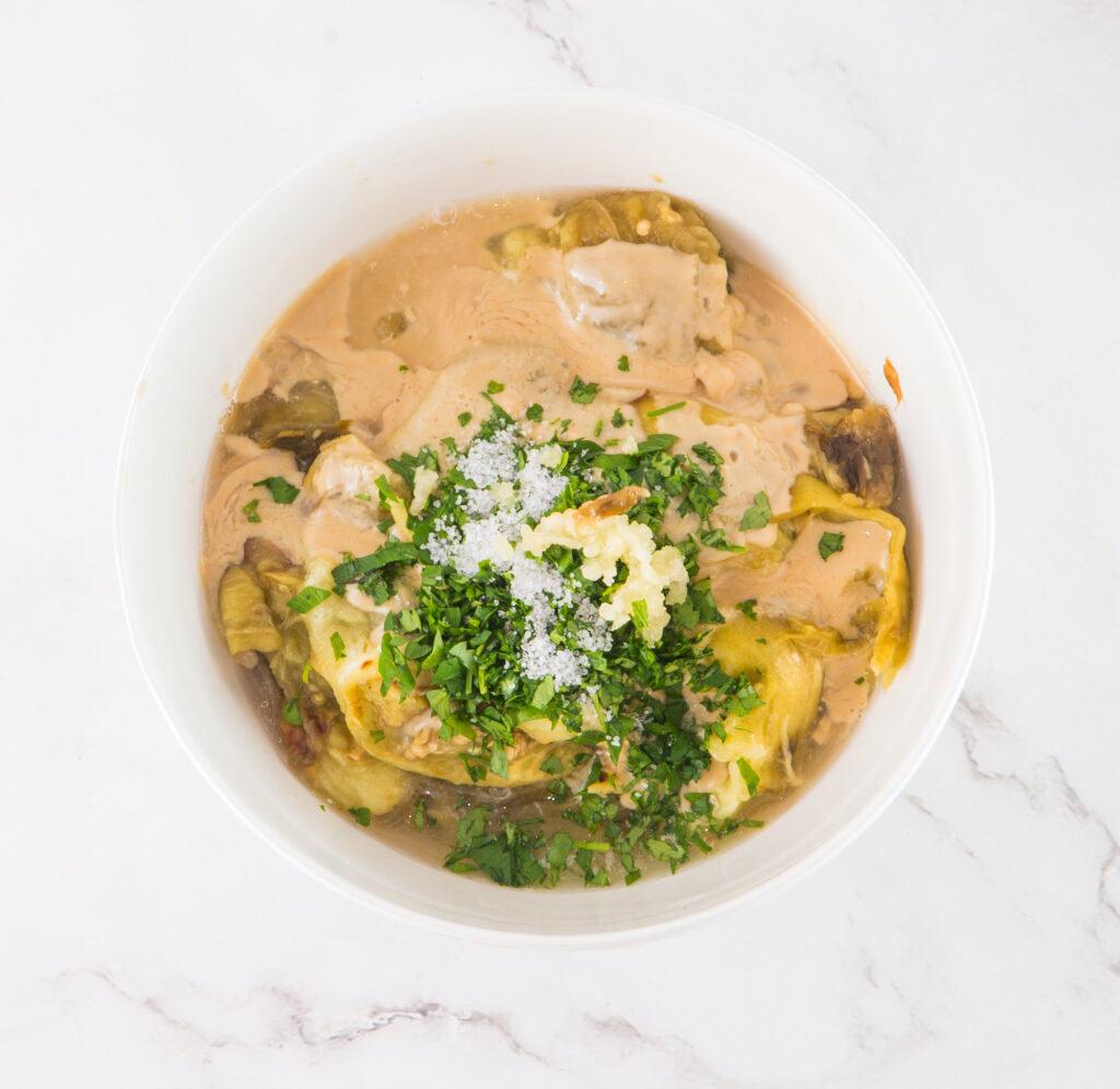 A white bowl filled with creamy artichoke dip, topped with chopped parsley and grated cheese, on a marble surface.