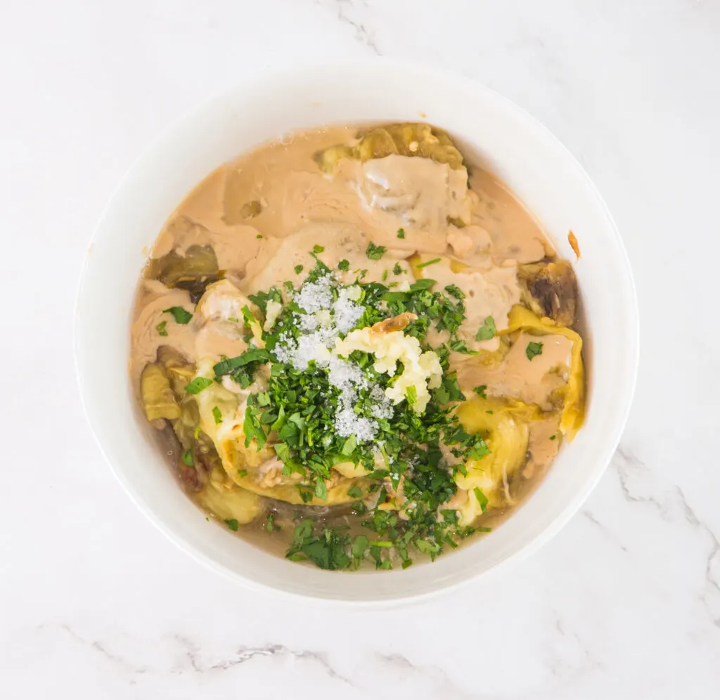 A white bowl filled with creamy artichoke dip, topped with chopped parsley and grated cheese, on a marble surface.