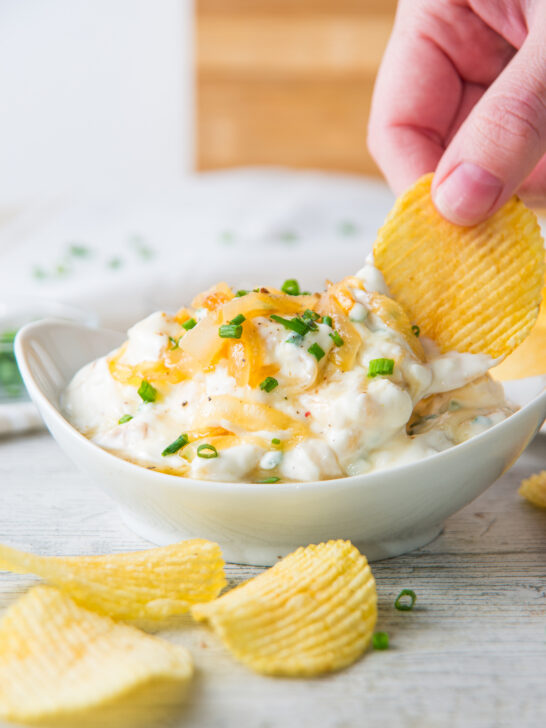 A hand dips a potato chip into a bowl of French onion dip, its creamy surface sprinkled with chopped green onions, on a light wooden surface surrounded by scattered chips.