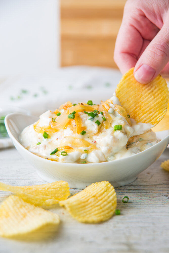 A hand dips a potato chip into a bowl of French onion dip, its creamy surface sprinkled with chopped green onions, on a light wooden surface surrounded by scattered chips.