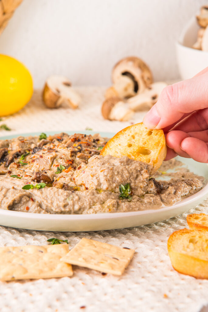 A hand dips a toasted bread slice into a bowl of mushroom dip garnished with herbs. Nearby are crackers and whole mushrooms.