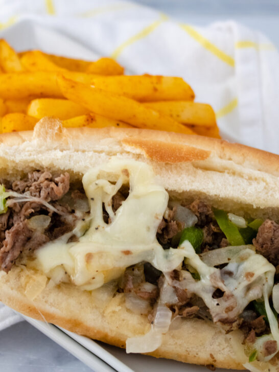 Philly cheesesteak sandwich on white plate with fries in background