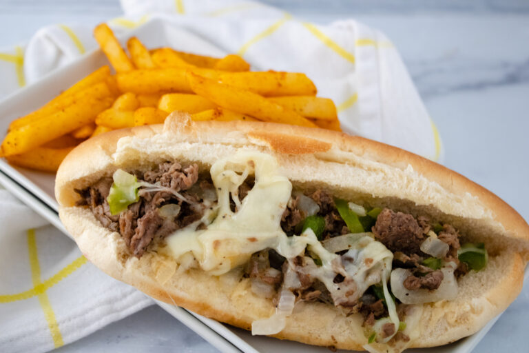 Philly cheesesteak sandwich on white plate with fries in background