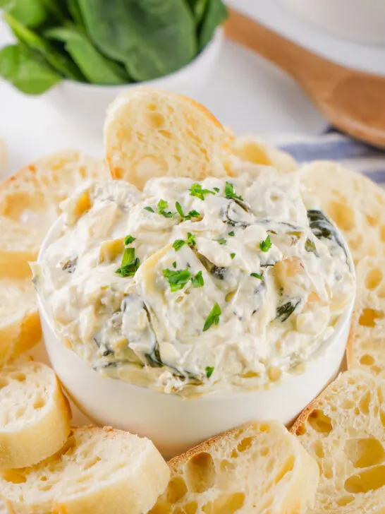 A bowl of creamy spinach artichoke dip is surrounded by slices of toasted bread.