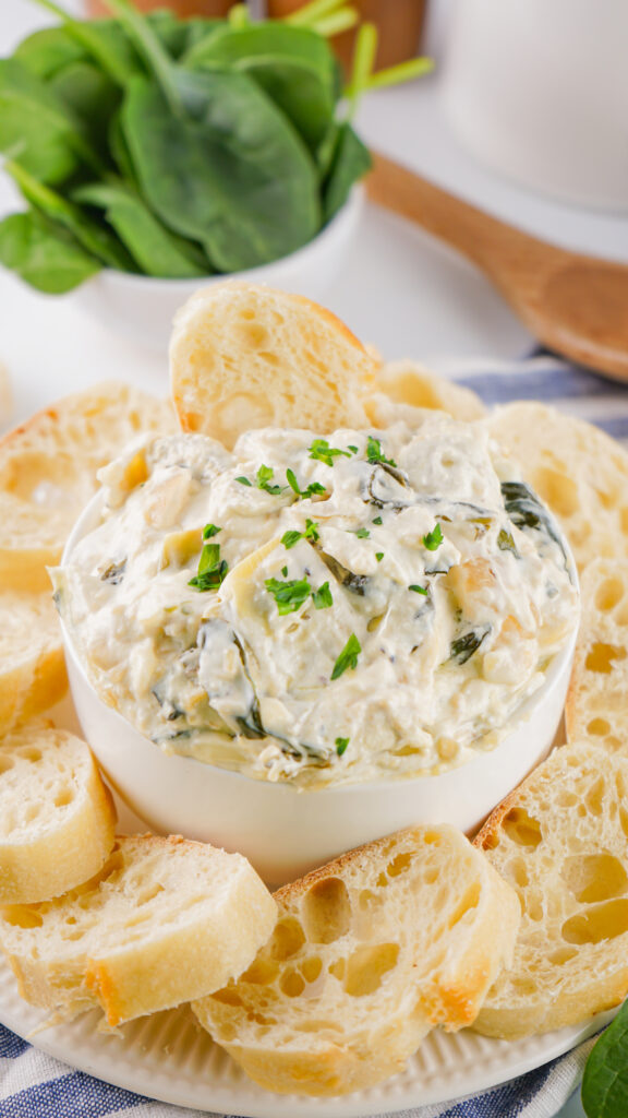 A bowl of creamy spinach artichoke dip is surrounded by slices of toasted bread.