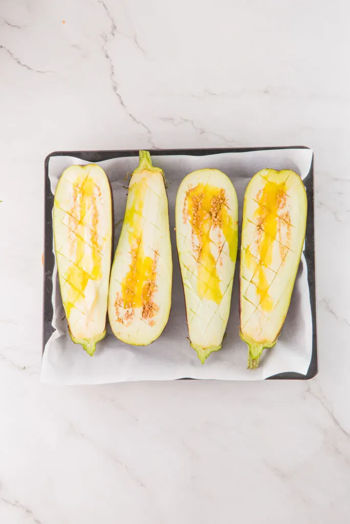 Four halved and scored eggplants on a baking sheet, drizzled with oil, ready for cooking.