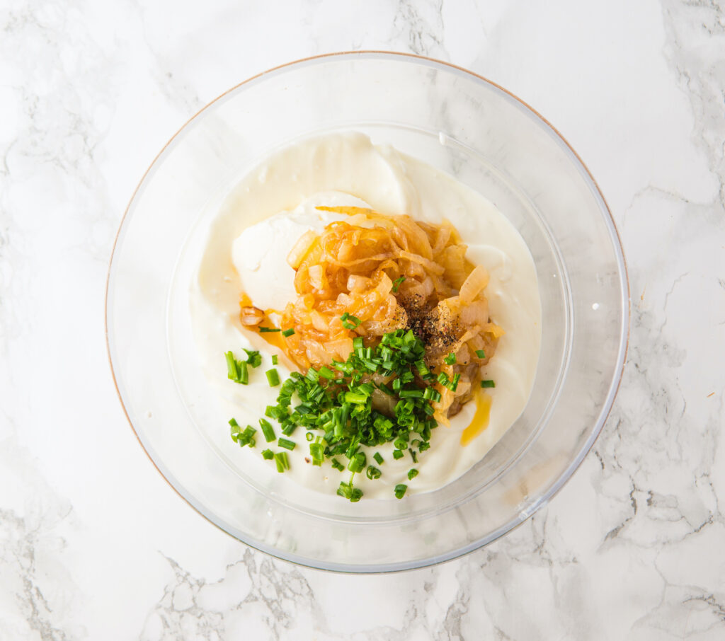 A glass bowl of creamy white yogurt, reminiscent of a French onion dip, is decadently topped with caramelized onions, chopped chives, and a sprinkle of black pepper, elegantly placed on a marble surface.