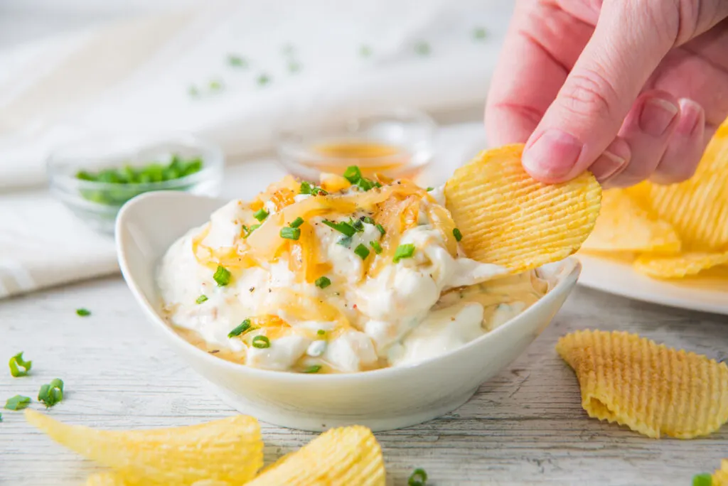 A hand dips a ridged potato chip into a bowl of creamy French onion dip, garnished with chives. Crinkle-cut chips are scattered around the bowl.