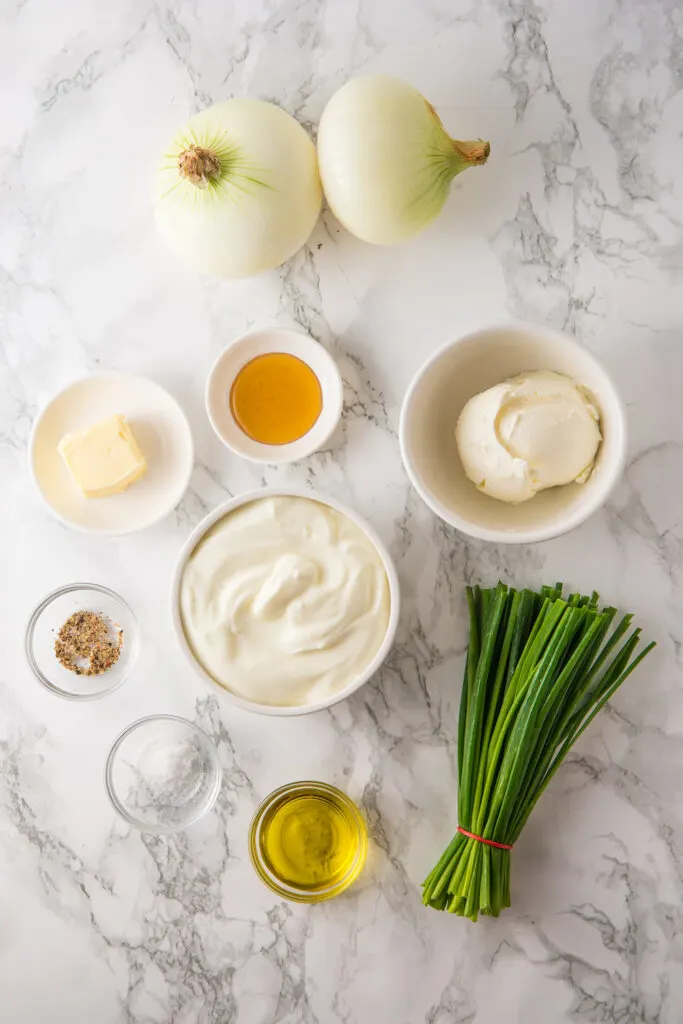 Ingredients for a French onion dip recipe arranged on a marble surface: white onions, sour cream, cream cheese, chives, butter, olive oil, honey, salt, and pepper.