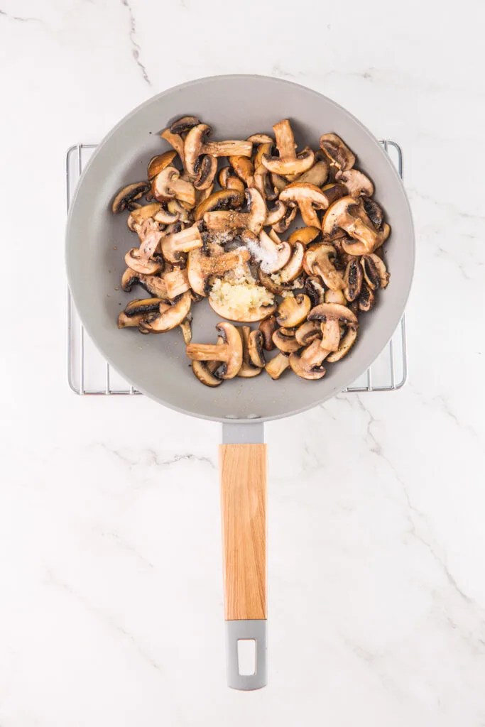 A frying pan with sautéed mushrooms, garlic, and seasonings rests on a cooling rack atop a marble countertop, ready to inspire the perfect mushroom hummus.