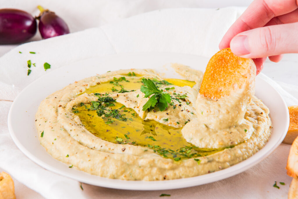 A hand dips a chip into a plate of hummus garnished with olive oil and herbs.