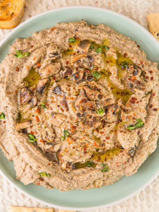 A plate of creamy mushroom hummus, elegantly garnished with a drizzle of oil, fresh herbs, and a sprinkle of chili flakes, is beautifully surrounded by rustic bread slices and crispy crackers.